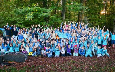Die Mitglieder der Pfadi Adler Aarau posieren mit vielen Kindern und Jugendlichen in blauer Pfadikleidung und erhobenen Händen für ein Gruppenfoto im Wald in Aarau (5000, Kanton Aargau).