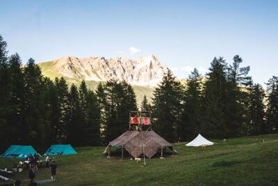 In einem alpinen Zeltlager der Pfadi Huenenberg in Huenenberg (6331, Kanton Zug) stehen mehrere Zelte vor dichtem Wald mit einer markanten Bergkette im Hintergrund.