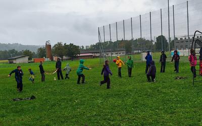 Auf einer Wiese in Toffen (3125, Kanton Bern) spielen Kinder bei feuchtem Wetter gemeinsam Ball und bewegen sich in einer Gruppe im Freien.