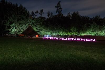 Bei Nacht liegt auf einer Wiese in Uetendorf (3661, Kanton Bern) ein beleuchteter Schriftzug vor Zelten und angestrahltem Waldrand.