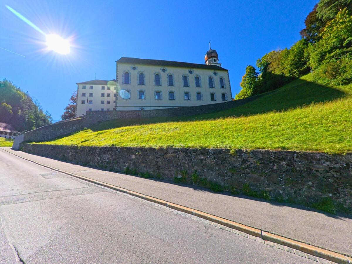 Auf einem sonnigen Hang in Pfäfers (7312, Kanton St. Gallen) steht ein barockes Gebäude mit Turmkuppel oberhalb einer steinernen Mauer und einer Strasse.