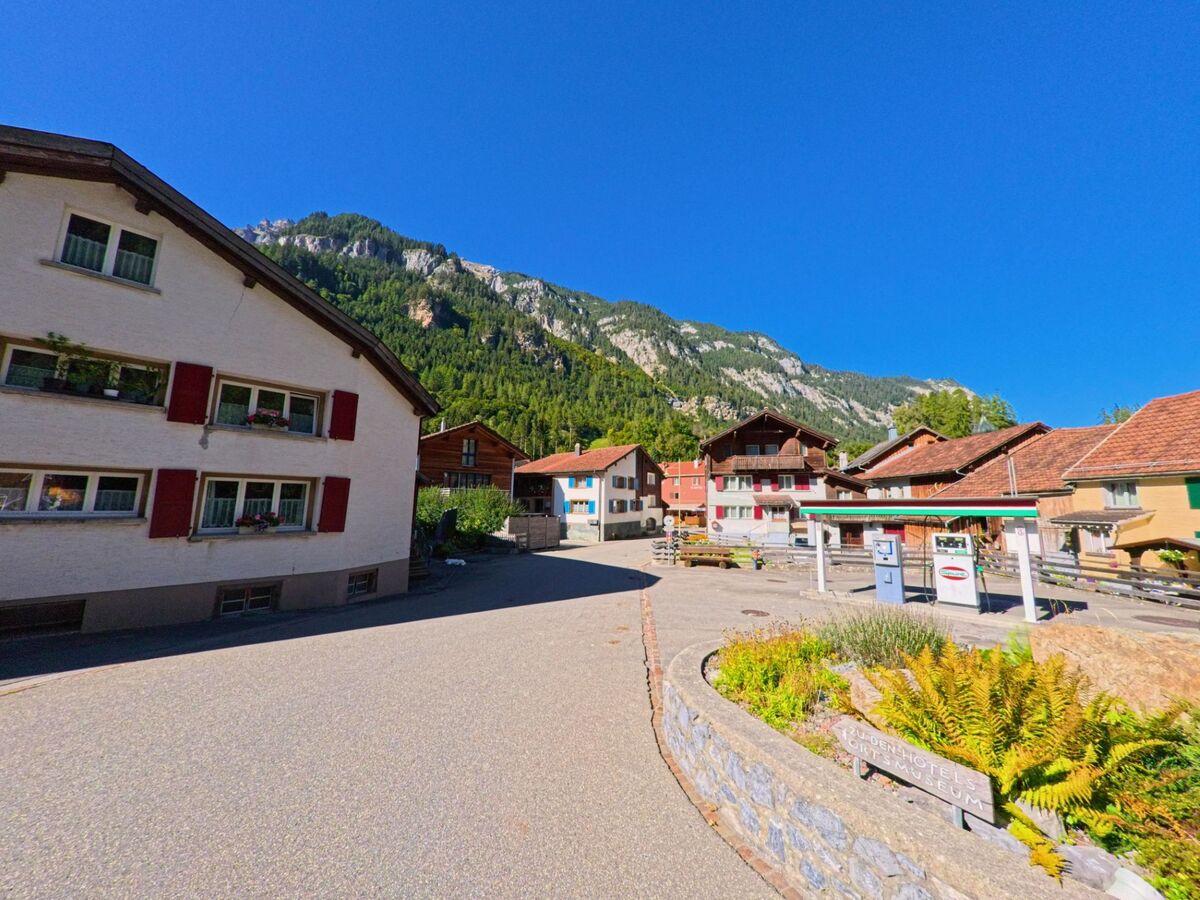 Im Dorfzentrum von Pfäfers (7315, Kanton St. Gallen) stehen traditionelle Häuser mit roten Fensterläden vor steilen Felswänden und einem klaren blauen Himmel.