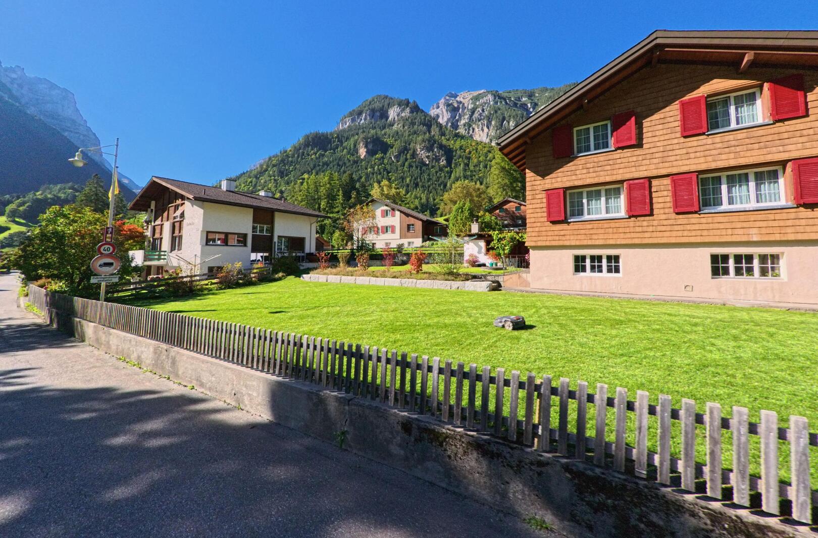 Vor den steilen Berghängen von Pfäfers (7315, Kanton St. Gallen) stehen gepflegte Wohnhäuser mit roten Fensterläden und blühenden Gärten unter klarem Himmel.