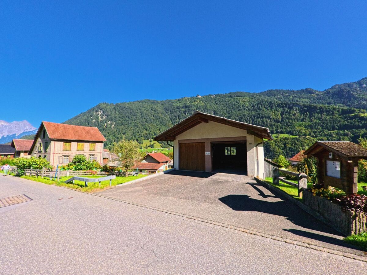 Ein kleines Gebäude mit Holztor steht in Pfäfers (7317, Kanton St. Gallen) an einer ruhigen Strasse mit Blick auf bewaldete Hügel und rote Ziegeldächer.