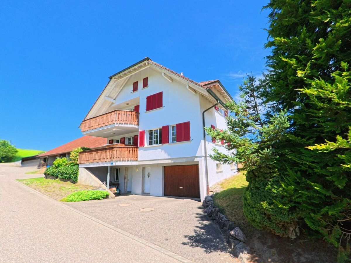 Ein weisses Wohnhaus mit roten Fensterlaeden und Holzbalkonen steht in Pfaffnau (PLZ 6264, Kanton Luzern) an einer ruhigen Nebenstrasse, flankiert von dichtem Gruen und Nachbargebaeuden.