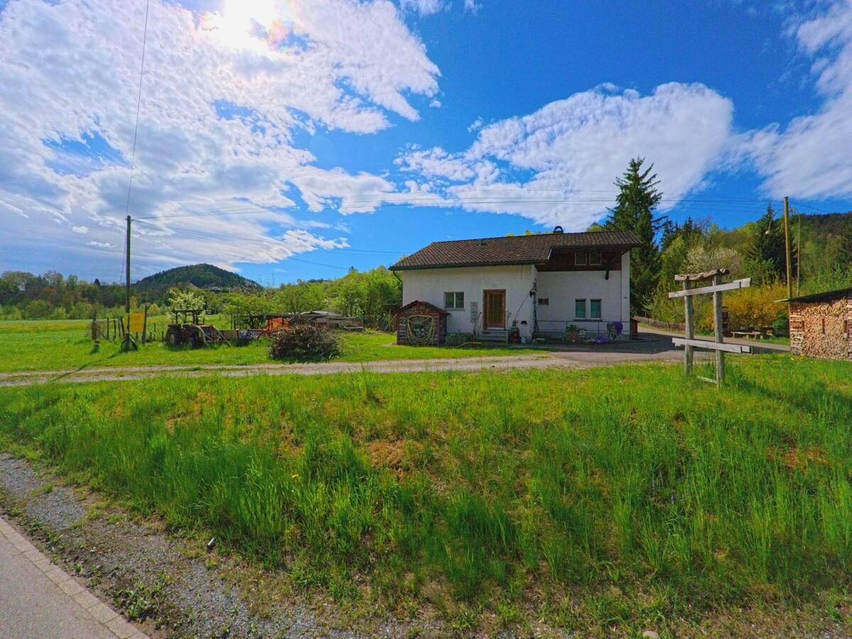 In Pfungen im PLZ-Bereich 8422 im Kanton Zürich steht ein einzelnes Bauernhaus mit Holzschuppen am Rand einer grünen Wiese mit Blick auf bewaldete Hügel.