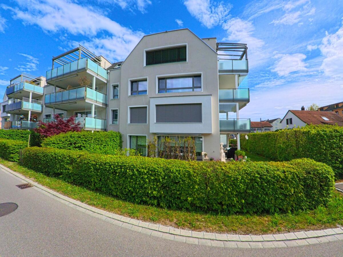In Pfungen im Kanton Zürich, PLZ 8422, steht ein modernes Mehrfamilienhaus mit grossen Balkonen und gepflegter Hecke an einer ruhigen Quartierstrasse.