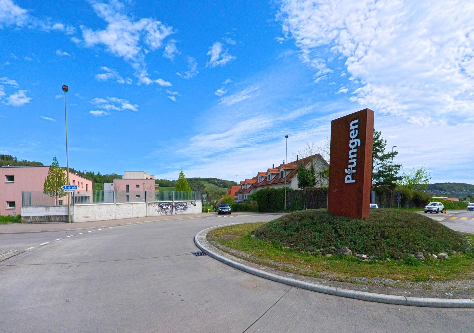 Am Ortseingang von Pfungen (8422, Kanton Zürich) steht eine rostfarbene Stele mit Ortsname, umgeben von Wohnhäusern und einem Kunstgraffito an der Betonmauer.