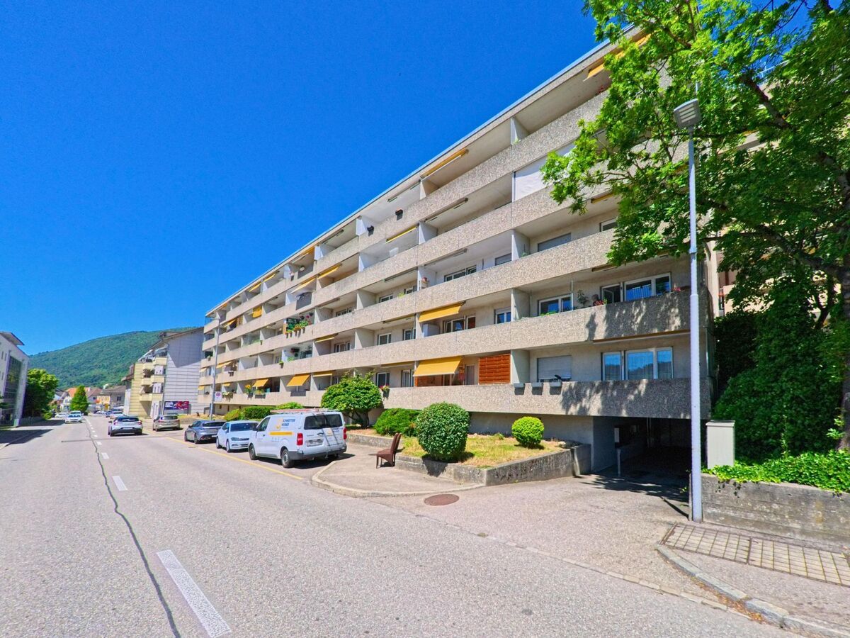 In Pieterlen (2542, Kanton Bern) erstreckt sich ein langgezogenes Wohngebäude mit Balkonen und gelben Markisen entlang einer vielbefahrenen Strasse.