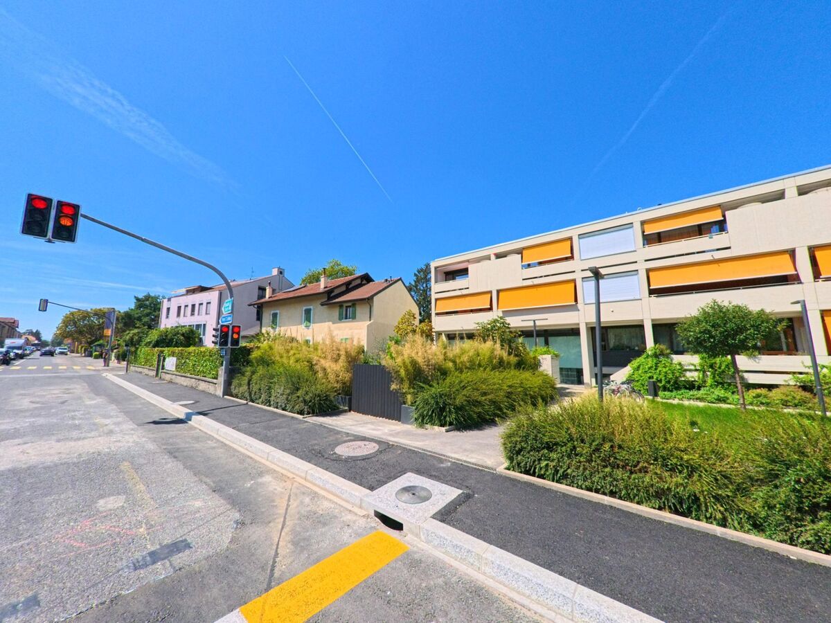 An einer Kreuzung in Plan les Ouates (1228, Kanton Genf) stehen moderne Wohngebaeude mit orangen Sonnenstoren neben aelteren Hausern.