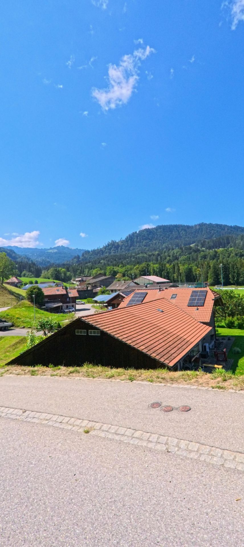 Von einer ansteigenden Strasse in Plasselb (1737, Kanton Freiburg) blickt man auf Hauser mit roten Daechern und Solarpanels, umgeben von gruener Landschaft und bewaldeten Huegeln.