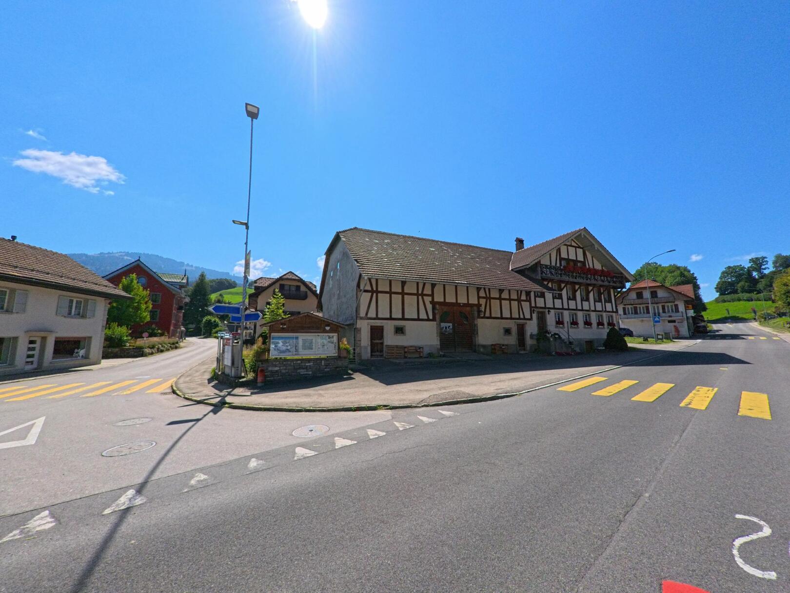 An der Hauptstrasse in Plasselb (1737, Kanton Freiburg) steht ein traditionelles Fachwerkhaus mit Blumenbalkon unter klarem Sommerhimmel, umgeben von weiteren Dorfhäusern und Zebrastreifen.