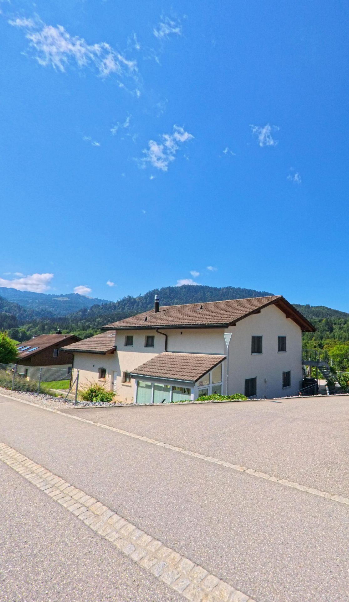 Ein Einfamilienhaus mit braunem Dach steht an einer ansteigenden Strasse in Plasselb (1737, Kanton Freiburg), umgeben von gruener Landschaft und bewaldeten Huegeln im Hintergrund.