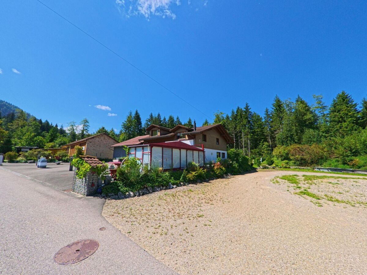 Am Waldrand von Plasselb (1737, Kanton Freiburg) steht ein Haus mit Wintergarten und Gartenanlage, daneben fuehrt eine schmale Strasse an mehreren Gebaeuden vorbei.