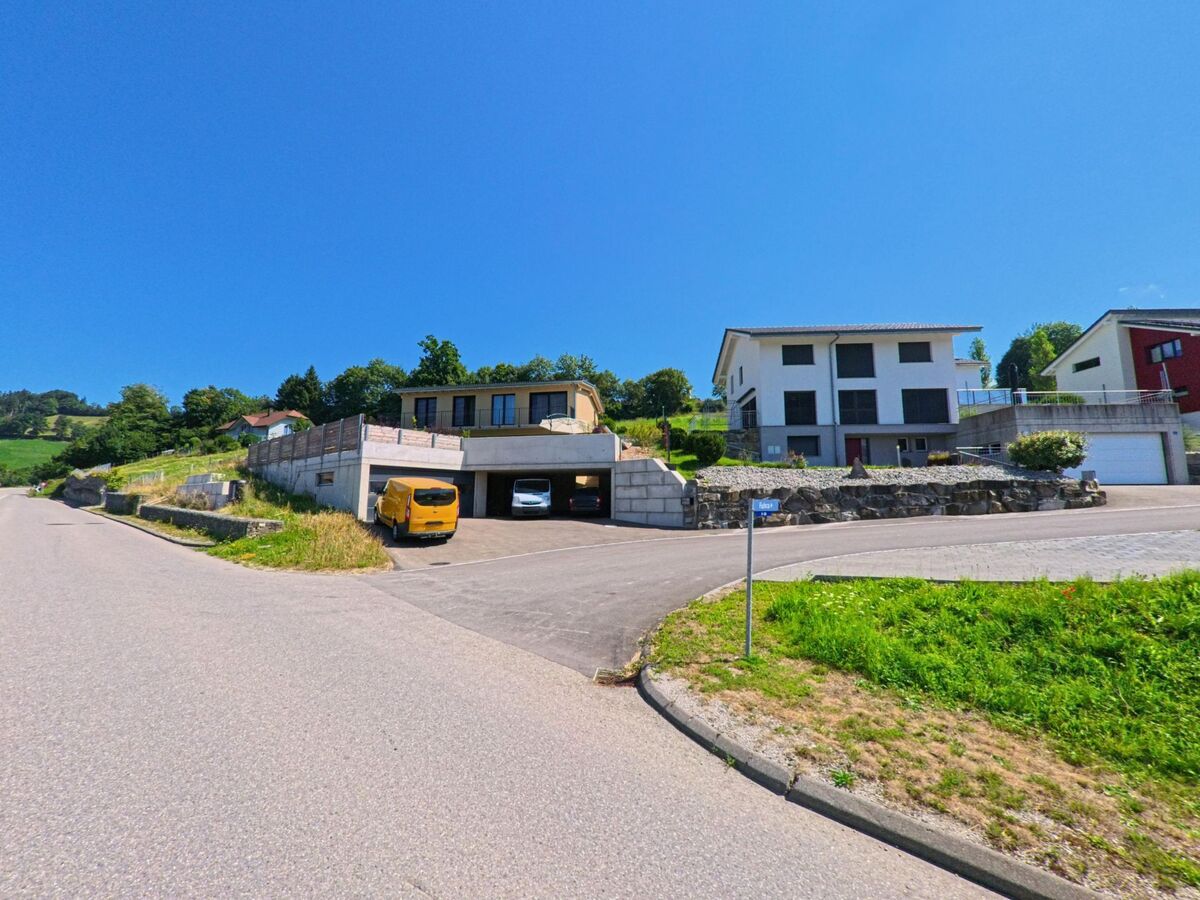 In einem Wohnquartier von Plasselb (1737, Kanton Freiburg) stehen moderne Hauser mit Garagenanlagen an einer leicht ansteigenden Strasse unter strahlend blauem Himmel.