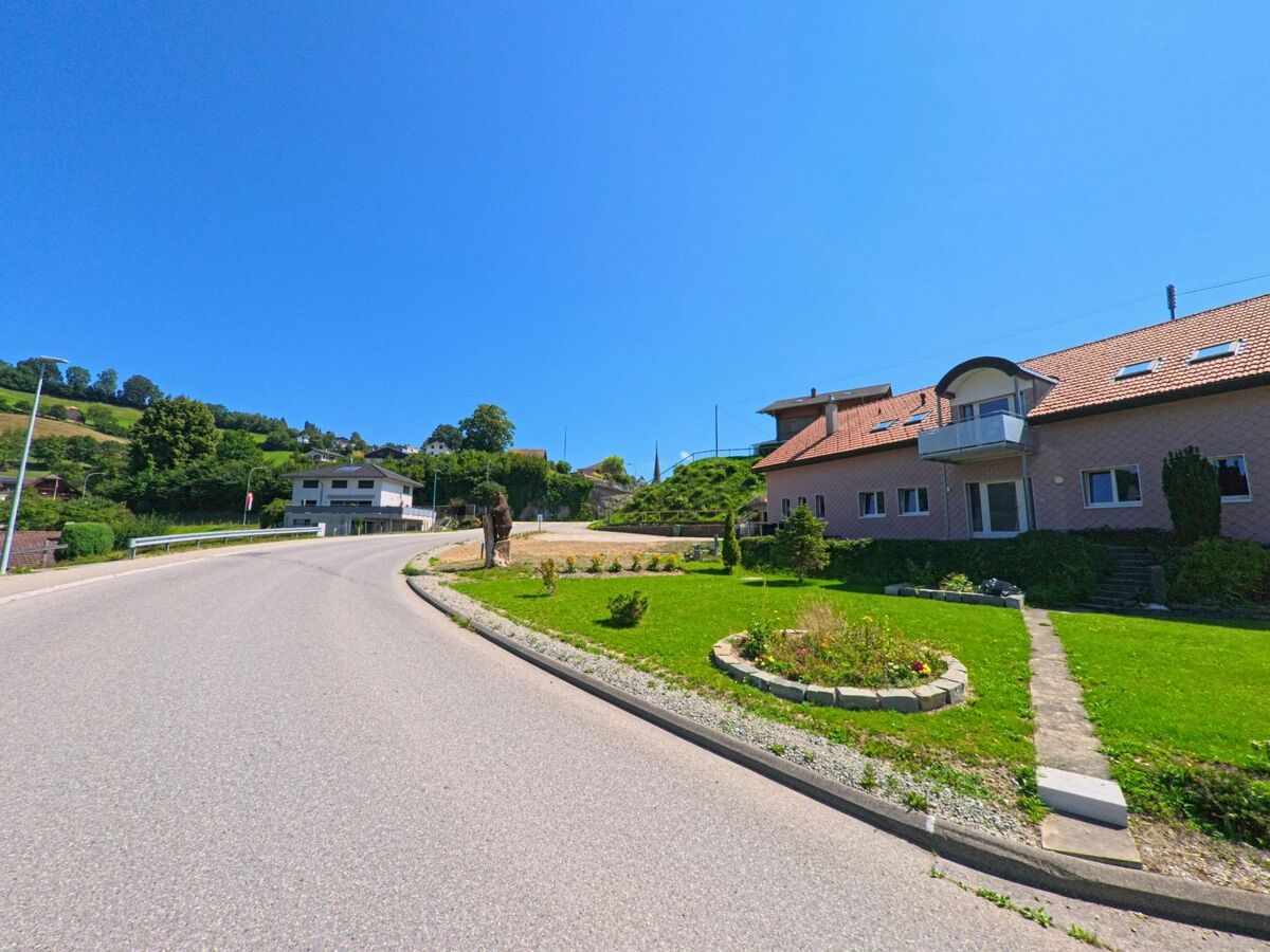 Eine leicht geschwungene Strasse fuehrt durch ein Wohnquartier in Plasselb (1737, Kanton Freiburg), vorbei an gepflegten Gaerten und Hausern mit roten Daechern unter blauem Himmel.