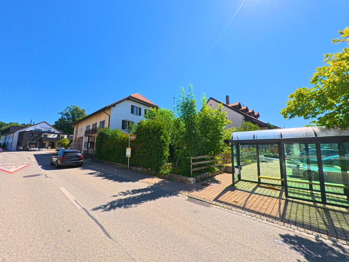 Eine Bushaltestelle mit Glasunterstand befindet sich in Plateau de Diesse (2515, Kanton Bern) an einer Strasse mit Wohnhaeusern und Baeumen im Hintergrund.