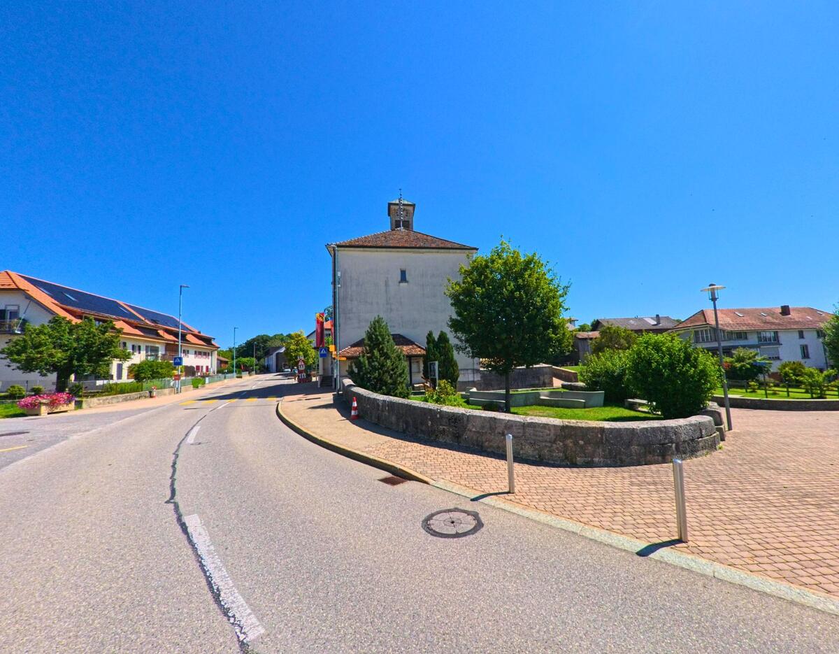 An einer sanft geschwungenen Strasse in Plateau de Diesse (2515, Kanton Bern) steht ein Gebaeude mit kleinem Turm neben einer gruengepflegten Platzanlage.