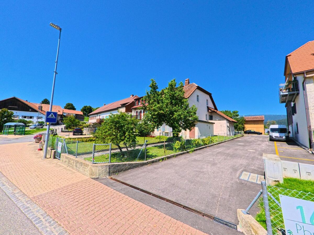In Plateau de Diesse (2515, Kanton Bern) stehen entlang einer Strasse mit Fussgaengerueberweg Wohnhaeuser mit roten Ziegeldach und gepflegten Gaerten.