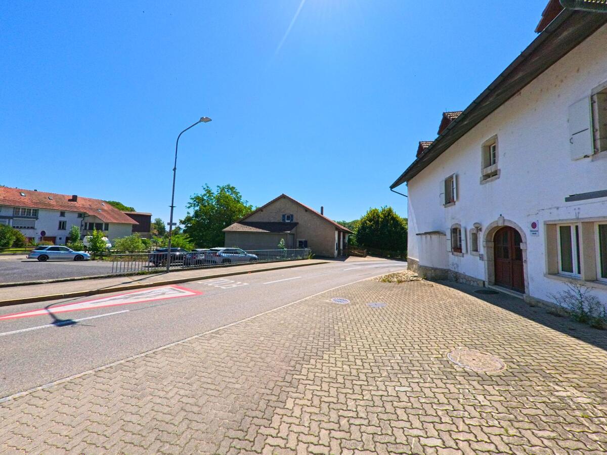 Ein weisses Wohnhaus mit historischen Fenstern und Holzportael befindet sich an einer Strasse in Plateau de Diesse (2515, Kanton Bern) neben einem kleinen Parkplatz.