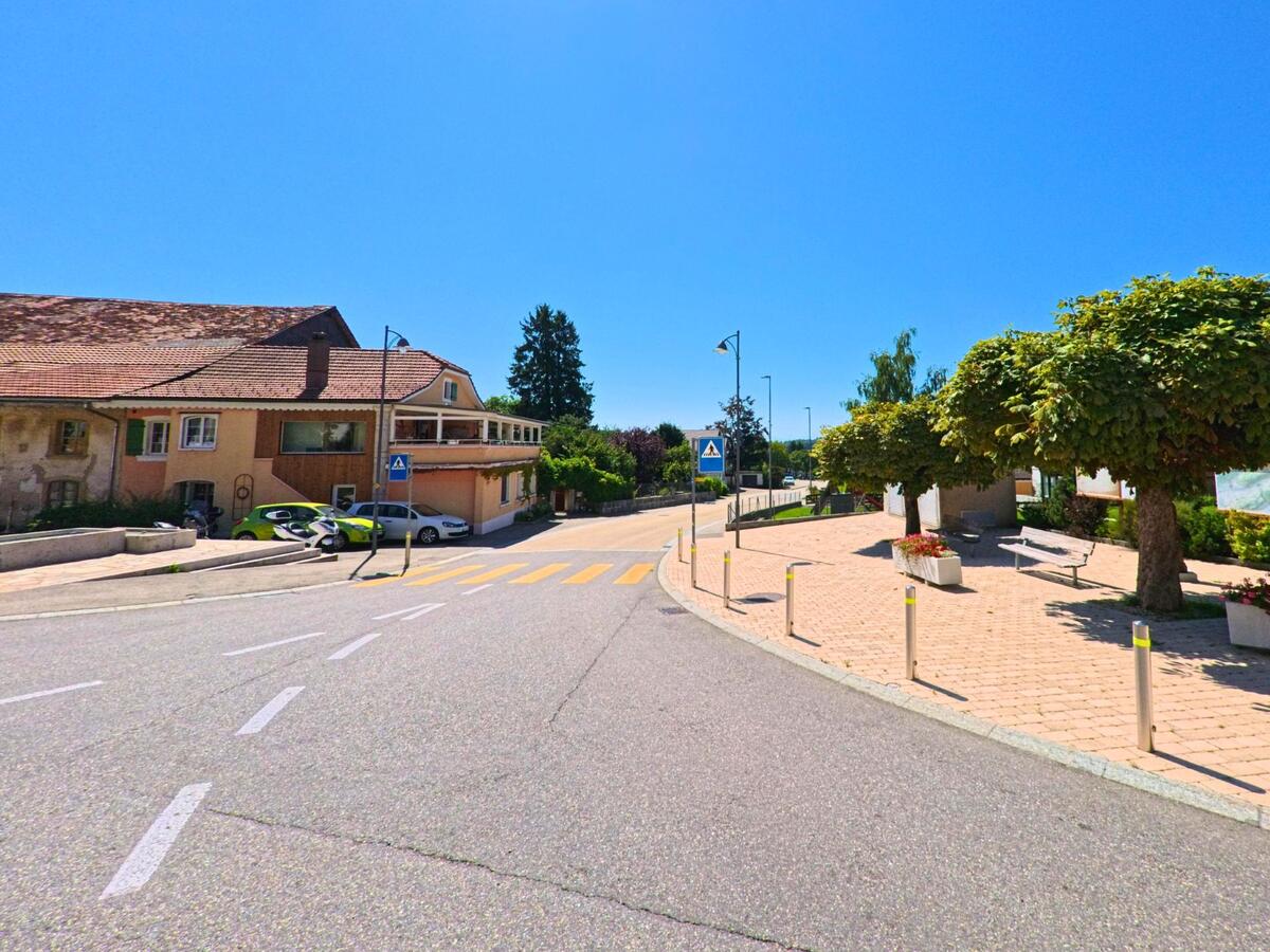 Ein Fussgaengerueberweg fuehrt in Plateau de Diesse (2516, Kanton Bern) an einer kleinen Platzanlage mit Schatten spendenden Baeumen und Sitzbaenken vorbei.