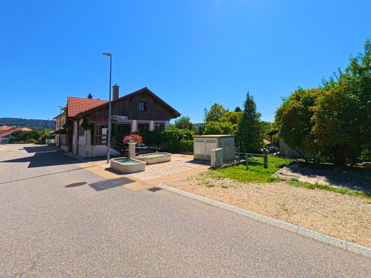 An einer sonnigen Strasse in Plateau de Diesse (2516, Kanton Bern) steht ein Haus mit rotem Ziegeldach und Holzelementen neben einem Brunnen mit Blumenschmuck.