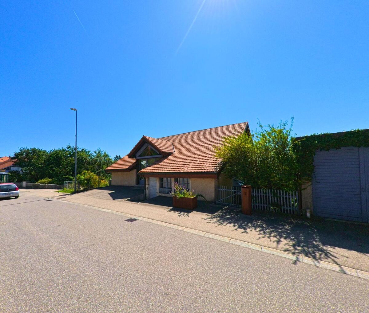Ein Haus mit rotem Ziegeldach und grossen Dachfenstern steht an einer ruhigen Strasse in Plateau de Diesse (2516, Kanton Bern) unter klarem Sommerhimmel.