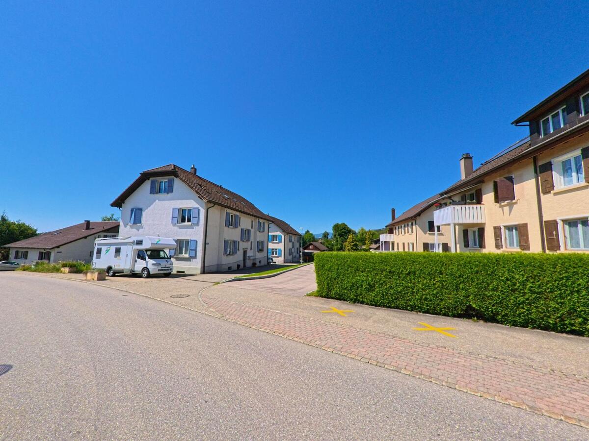 Mehrfamilienhaeuser mit gepflegten Fassaden und Fensterlaeden praegen eine ruhige Wohnstrasse in Plateau de Diesse (2516, Kanton Bern) unter blauem Himmel.