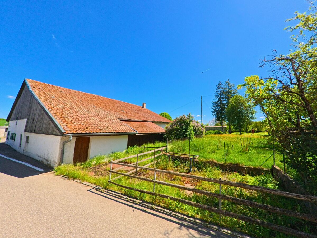In Pleigne (2807, Kanton Jura) steht ein landwirtschaftliches Gebaeude mit rotem Ziegeldach neben einer umzaeunten Gruenflaeche an einer schmalen Strasse.
