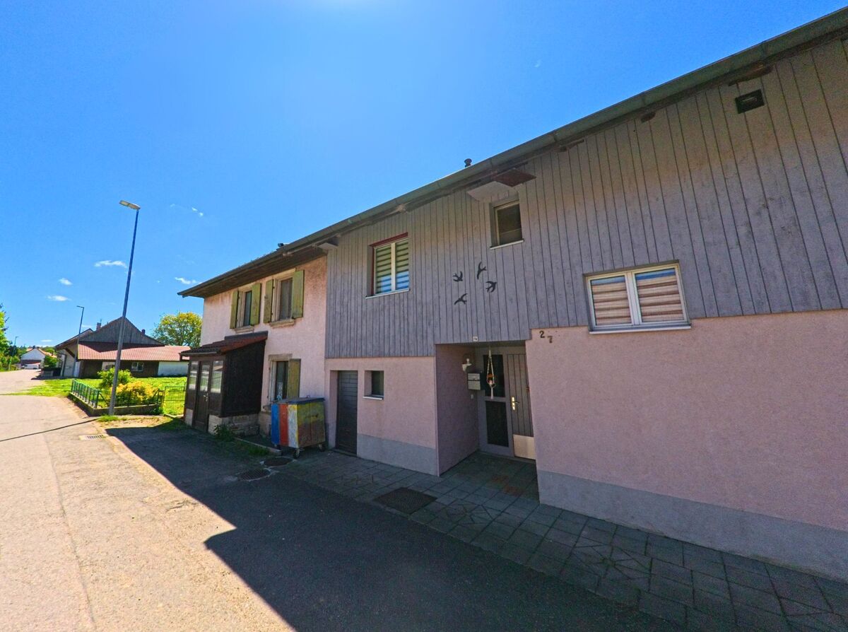 In Pleigne (2807, Kanton Jura) steht ein schlichtes Wohnhaus mit Holzfassade und kleinen Fenstern direkt an einer schmalen Strasse mit angrenzendem Gruenbereich.