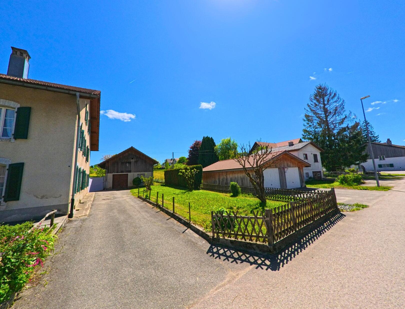 In Pleigne (2807, Kanton Jura) liegen mehrere Wohnhaeuser und Nebengebaeude entlang einer ruhigen Strasse mit kleinen Gaerten und Einfahrten.