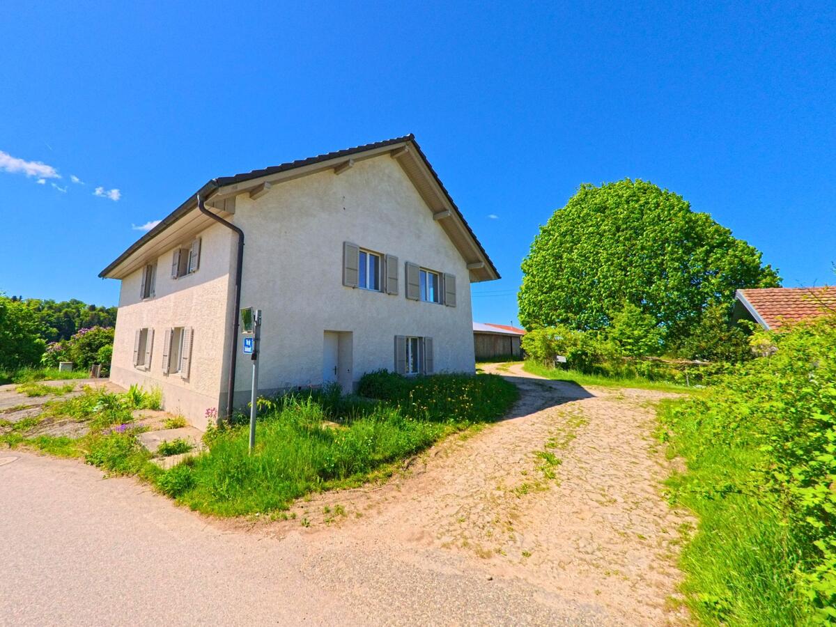 In Pleigne (2807, Kanton Jura) steht ein helles Wohnhaus mit geschlossenen Fensterlaeden an der Einmuendung eines unbefestigten Weges in ländlicher Umgebung.