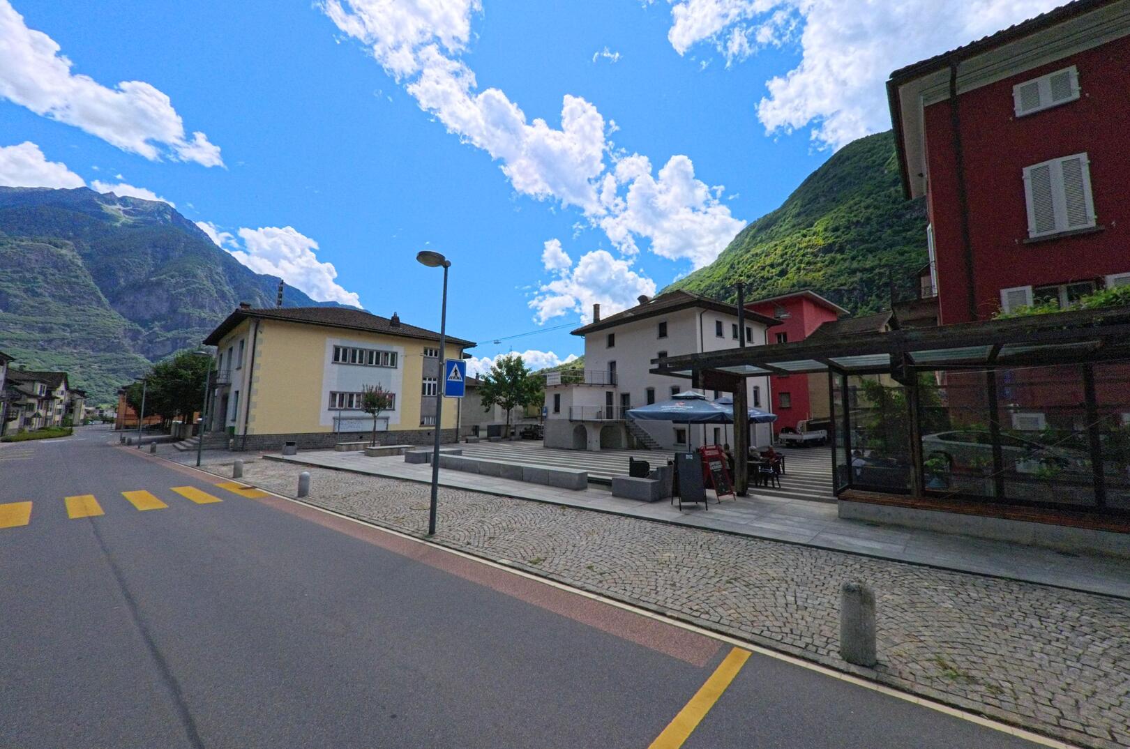 In Pollegio (6742, Kanton Tessin) führt eine gepflasterte Strasse mit Zebrastreifen durch das Dorfzentrum, umgeben von Wohnhäusern und Bergen im Hintergrund.