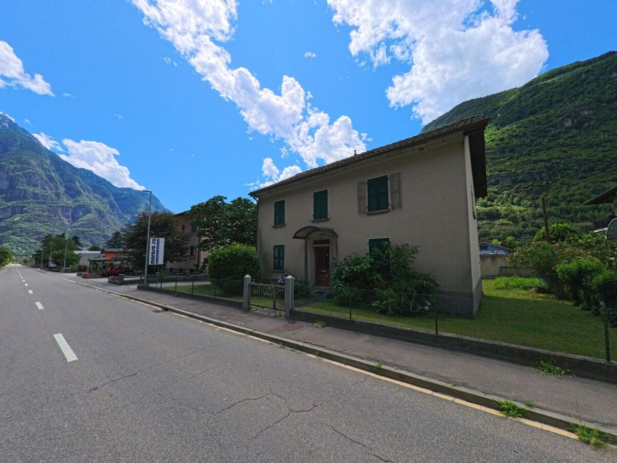 Ein Einfamilienhaus mit gruener Fensterlaeden steht in Pollegio (6742, Kanton Tessin) an einer ruhigen Strasse, eingerahmt von Garten und Berglandschaft.