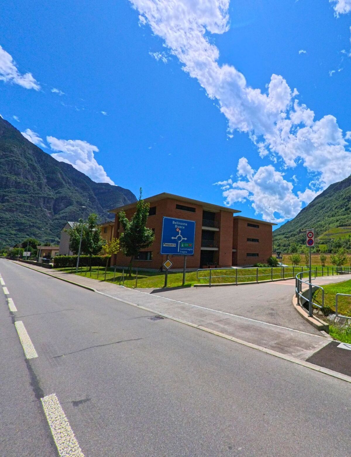 An der Hauptstrasse von Pollegio (6742, Kanton Tessin) steht ein modernes Backsteingebaeude mit Wegweiser in Richtung Bellinzona, umgeben von Bergen und blauem Himmel.
