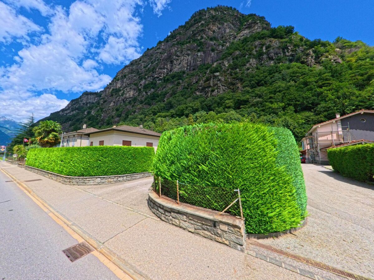 In Pollegio (6742, Kanton Tessin) saeumen gepflegte gruen geschnittene Hecken die Einfahrten von Wohnhaeusern am Fuss eines steilen bewaldeten Hanges.