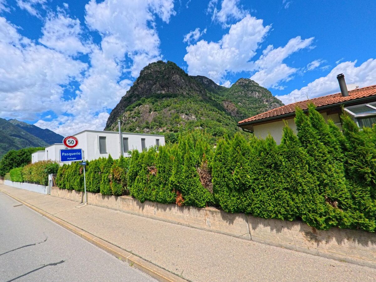 Am Ortseingang von Pollegio (6742, Kanton Tessin) steht ein Schild mit dem Quartiernamen Pasquerio, daneben moderne Wohnhaeuser vor einer steilen Felswand.