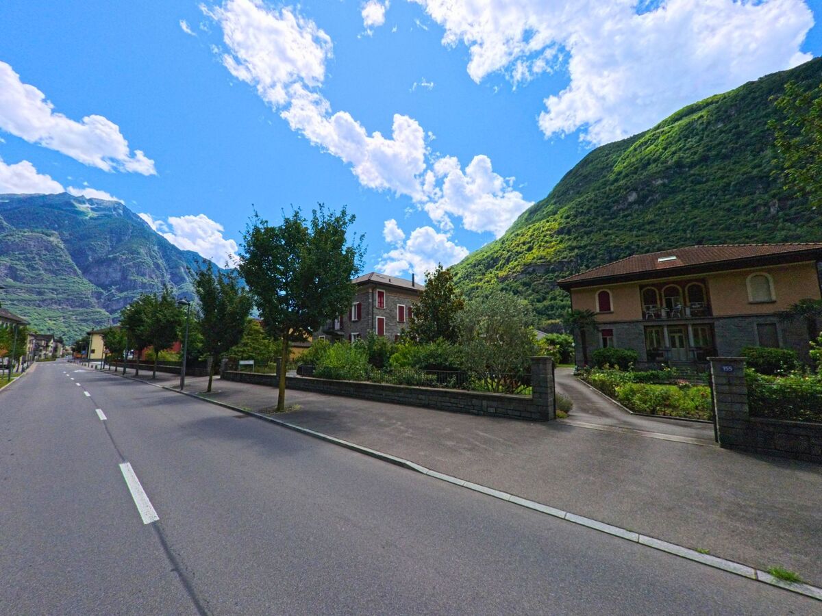 Entlang einer ruhigen Strasse in Pollegio (6742, Kanton Tessin) stehen gepflegte Wohnhaeuser mit Gaerten vor einer beeindruckenden Bergkulisse.