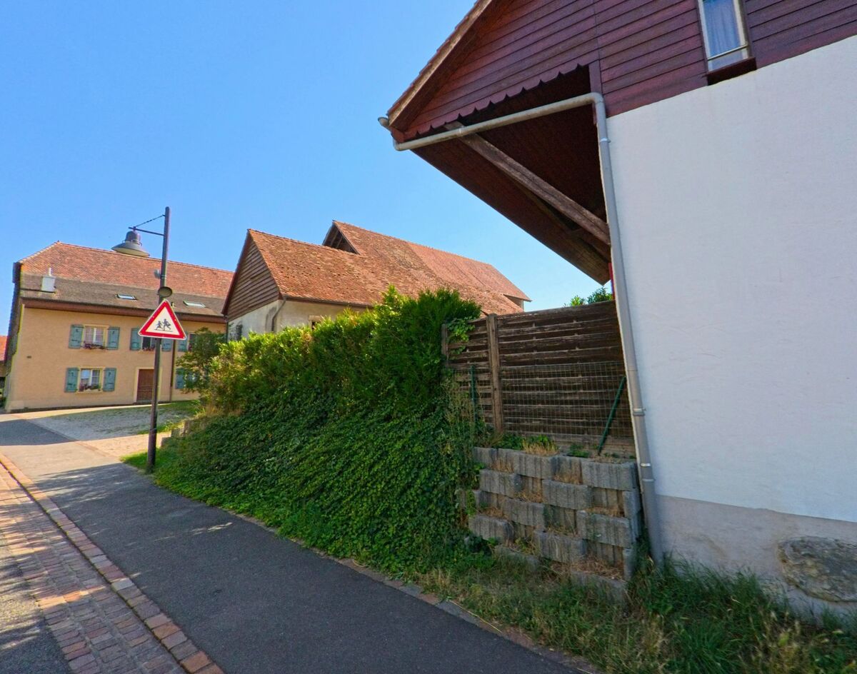 In Pomy (1405, Kanton Waadt) stehen traditionelle Dorfhaeuser mit roten Ziegeldächern entlang einer schmalen Strasse, neben der ein Fussgaengerschild angebracht ist.