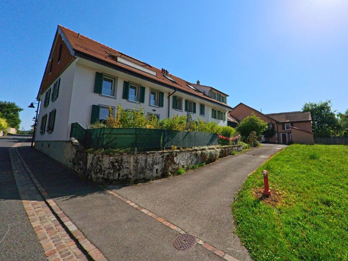 In Pomy (1405, Kanton Waadt) steht ein langgestrecktes Wohnhaus mit grünen Fensterläden an einer kleinen Strasse, daneben liegt eine Wiese mit Hydranten.