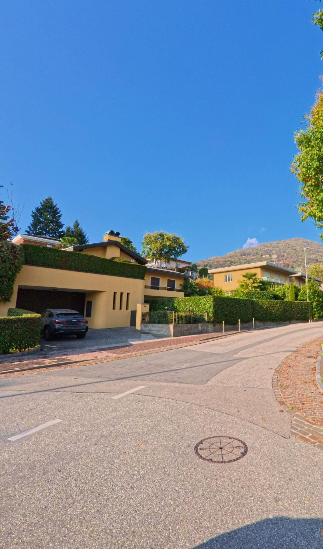 Ein modernes Einfamilienhaus mit gelber Fassade und gepflegter Hecke steht an einer ruhigen Strasse in Ponte Capriasca (6946, Kanton Tessin) mit Blick auf den bewaldeten Hügel im Hintergrund.