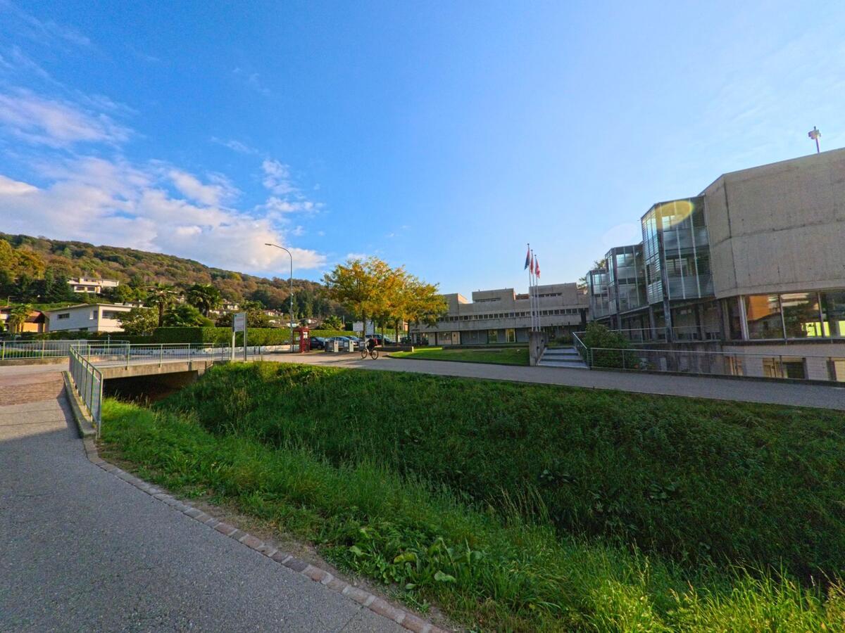 Vor dem modernen Schul- oder Verwaltungsgebäude in Ponte Capriasca (6946, Kanton Tessin) weht die Schweizer Flagge, während im Hintergrund bewaldete Hügel zu sehen sind.