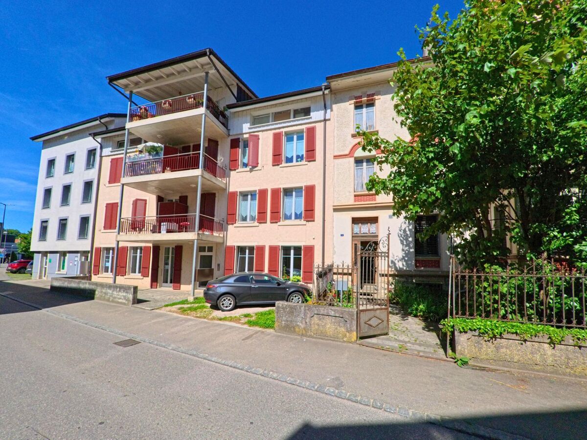 In einem Wohnquartier von Porrentruy (2900, Kanton Jura) stehen mehrere Mehrfamilienhäuser mit roten Fensterläden und Balkonen entlang einer schmalen Seitenstrasse.