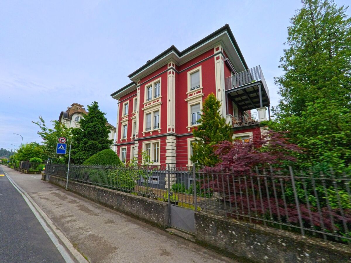 Eine rot gestrichene Stadtvilla mit klassizistischen Elementen steht in Porrentruy (2900, Kanton Jura) hinter einem schmiedeeisernen Zaun an einer ruhigen Strasse.