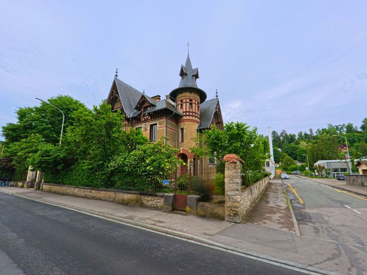 In Porrentruy (2900, Kanton Jura) erhebt sich eine burgähnliche Villa mit spitzen Türmen und reicher Fassadengestaltung hinter einer alten Steinmauer an einer Kreuzung.