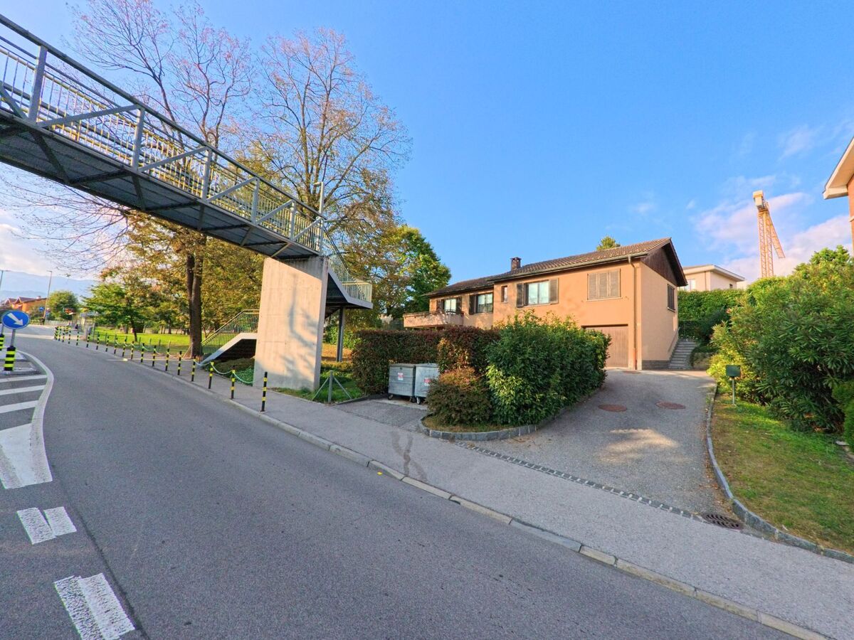 An einer Strasse in Porza (6948, Kanton Tessin) überspannt eine Fussgängerbrücke den Fahrraum, während daneben ein Wohnhaus mit Vorgarten und Gehweg liegt.