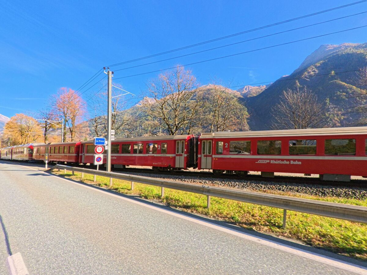 Ein roter Zug der Rhätischen Bahn fährt bei Poschiavo (7746, Kanton Graubünden) entlang der Strasse durch ein herbstliches Tal mit bewaldeten Berghängen.