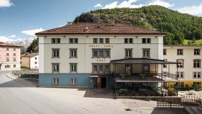 Das Post Hotel Loewe in Mulegns (7455, Kanton Graubuenden) zeigt seine historische Fassade mit gedeckter Terrasse vor alpiner Bergkulisse.