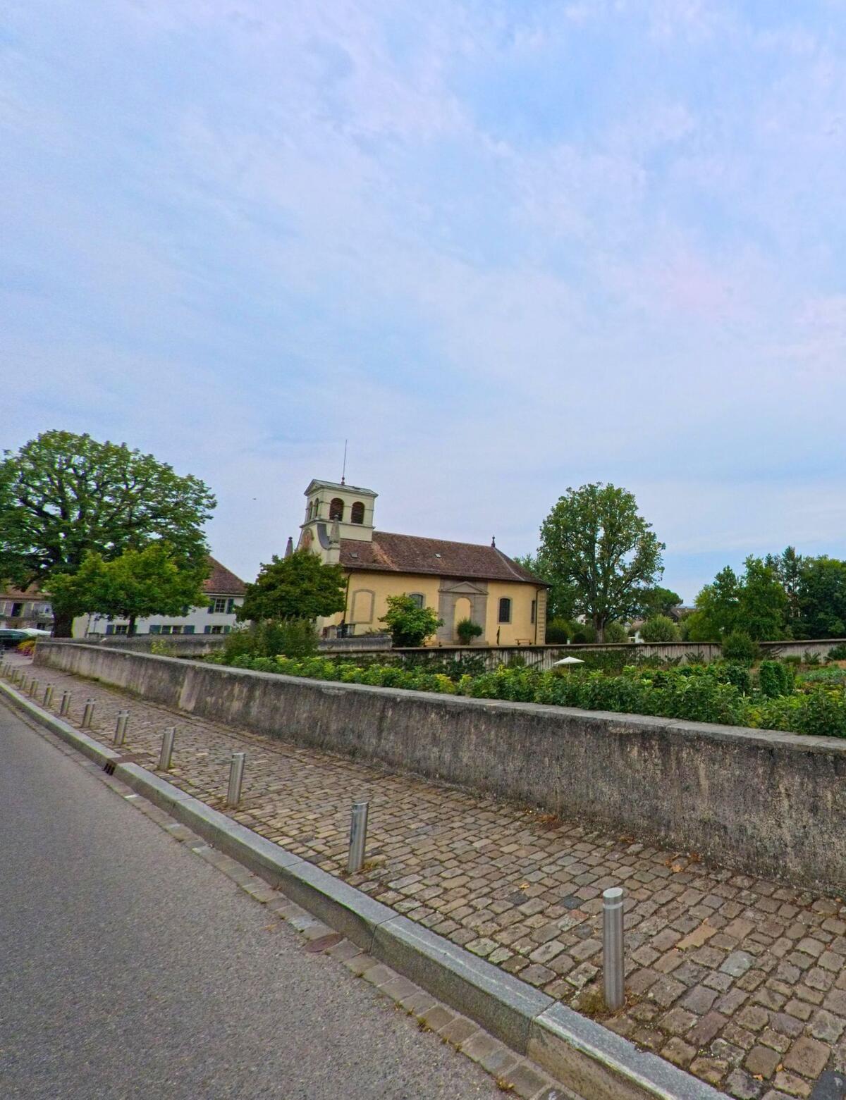 Eine Kirche mit hellem Turm und Ziegeldach erhebt sich in Prangins (1197, Kanton Waadt) hinter einer alten Mauer und umgeben von Bäumen.