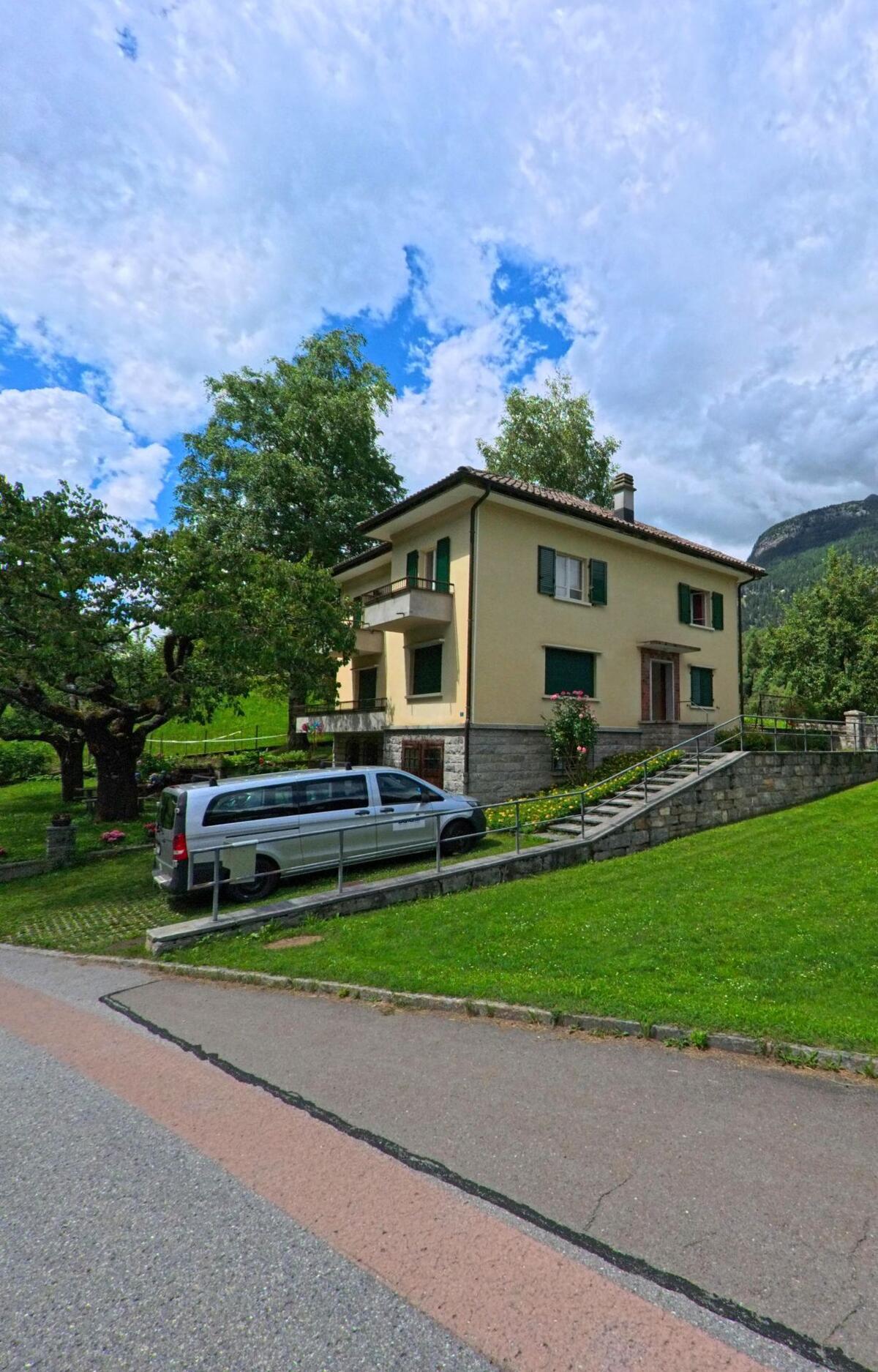 Ein gelbes Wohnhaus mit grünen Fensterläden steht in Prato (Leventina) (6773, Kanton Tessin) auf einem gepflegten Grundstück mit Rasen, Blumen und einer kleinen Auffahrt.